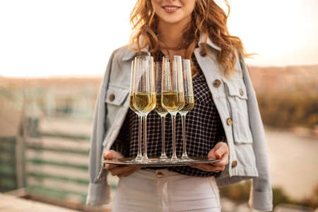 Young blonde smiling woman holding tray with champagne sparkling wine in flute glasses, standing outdoors over blurred countryside background, celebrating birthday party on cool summer eveningの写真素材