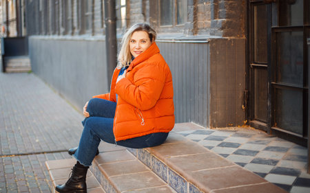 Happy smiling middle aged woman in orange down jacket sitting on concrete stairs outdoors and looking at camera with positive face expression, resting while walking in city on cold autumn dayの写真素材
