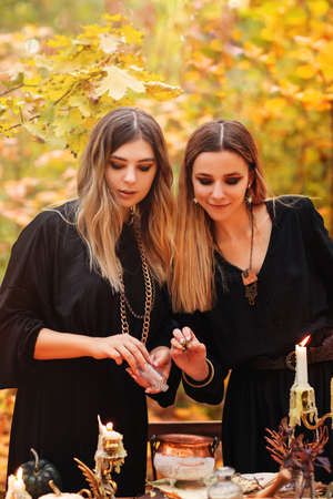 Young women in witch costumes sitting at table with magic ingredients on autumn day in forest