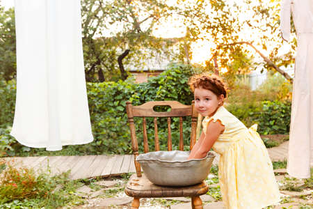 Cute little girl in dress washing white clothes in metal basin in backyard, child helping with chores, hanging laundry on clothesline and leaving it to dry in garden, drying clothes outdoors in windの写真素材