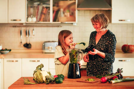 Loving grandma preparing smoothie with her cute little granddaughter while standing together in kitchen at home, blending various vegetables, preparing healthy and nutritious drink togetherの写真素材