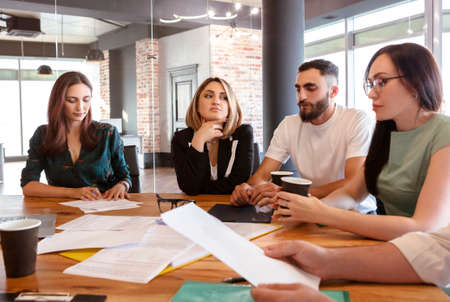 Creative team of diverse pensive young business people gathered for weekly meeting in office, sitting at big wooden desktop and discussing new business plan, group of four coworkers working togetherの写真素材