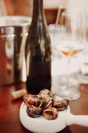 Vertical shot of escargots served on white ceramic pan alongside white wine, delicious cooked snails, enjoying traditional french cuisine in restaurant, selective focus on french dishの写真素材