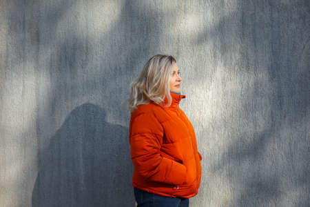 Happy positive middle-aged woman in orange down jacket smiling at camera while posing against...