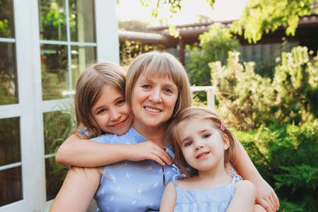 Happy loving middle-aged grandma hugging granddaughters while standing in garden on summer day,...