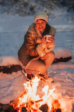 Happy young woman full of calmness and pacification in warm knitted hat and jacket sits on wooden sled by bonfire with cup of hot tea in her hands, surrounded by night snow-covered winter forestの写真素材
