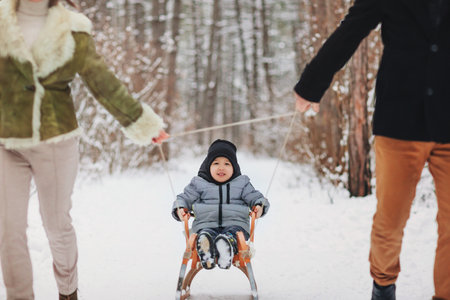 Young beautiful family of three walking in snowy forest, mom and dad dragging small son on sled, having fun on winter holiday outside. Seasonal vacation outdoor and spending leisure time with kidの写真素材