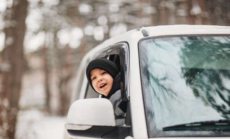 Excited little boy in black beanie and warm clothes sticking head out of car window, looking around while going through snowy forest in winter season. Happy childhoodの写真素材