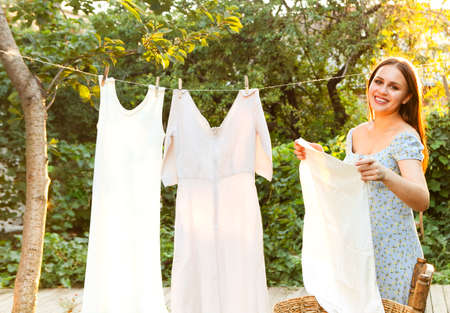 Young woman hanging laundry outdoors. Cute girl in dress washing white clothes in metal basin in...