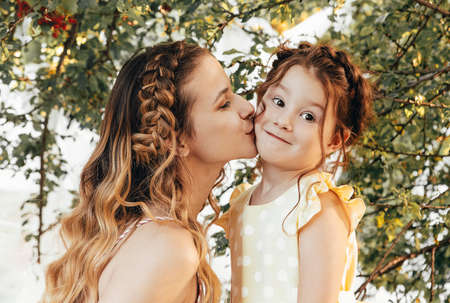 Maternity and loving motherhood. Beautiful young mom with braided hair kissing her lovely little sweet daughter in cheek while spending time together outdoors in garden under tree on summer dayの写真素材