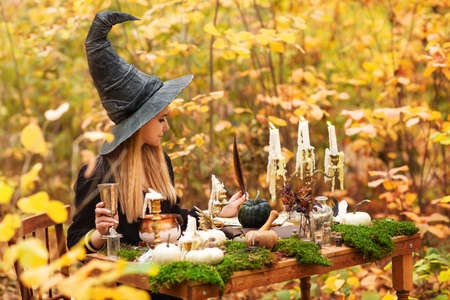 Young woman in witch costume sitting at table with magic ingredients and making notes on autumn day in forestの写真素材