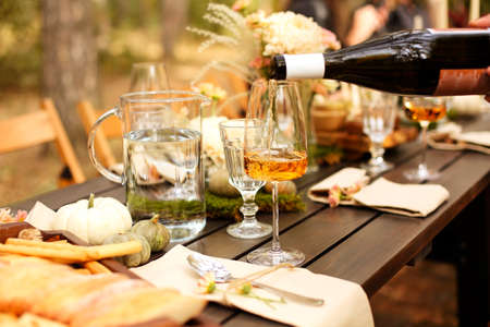 Glass of orange wine placed near dishware and bouquet of fresh flowers amidst assorted fruits on wooden table on autumn day in garden. Season party or celebrationの写真素材