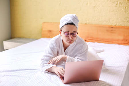 Happy woman in eyeglasses waiting and has video call on laptop lying on bed. Work from home conceptの写真素材