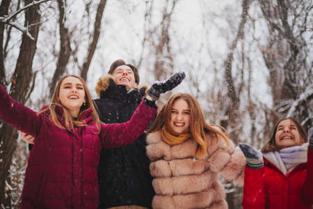 Group of four joyful best friends enjoying cold weather and first snow standing outdoor over snow covered trees, raising hands up and trying to catch falling snowflakes, looking up with happy smilesの写真素材