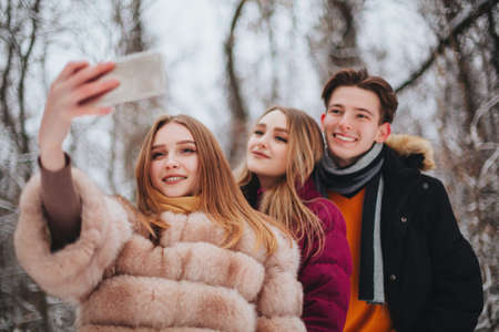 Image of three young cheerful happy best friends, two girls and boy making selfie on mobile phone smiling broadly on camera, standing against snow-covered trees and winter landscapeの写真素材