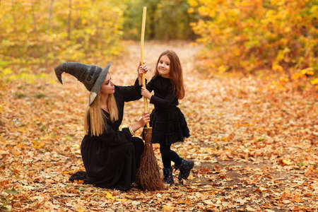 Full body woman and girl in witch costumes sweeping withered leaves from path with broom during Halloween celebration in autumn forestの写真素材