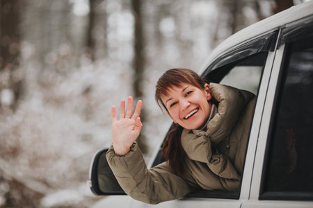 Cheerful female smiling and looking at camera while driving modern vehicle on blurred background of snowy countrysideの写真素材