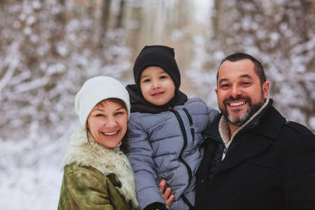 Cheerful parents walking behind glad boy with sleigh while spending time in snowy woods on winter dayの写真素材