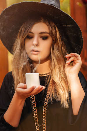 Young woman in Witch costume and hat looking mysteriously at candle in her hands isolated against autumn forest background with vintage wooden doors used as staged decoration for Halloween photo shootの写真素材