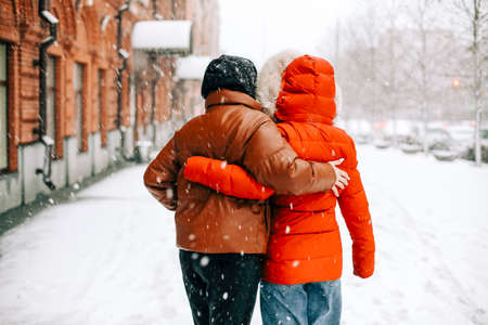 Back view of anonymous women in outerwear hugging each other and walking on snowy city street on winter dayの写真素材