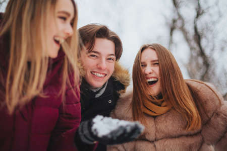 Group of four joyful best friends enjoying cold weather and first snow standing outdoor over snow covered trees, raising hands up and trying to catch falling snowflakes, looking up with happy smilesの写真素材