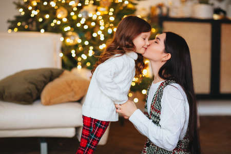 Beautiful loving mother gently kissing cute little daughter against festively decorated Christmas tree with glowing lights on background, happy family spending New Year eve at home togetherの写真素材