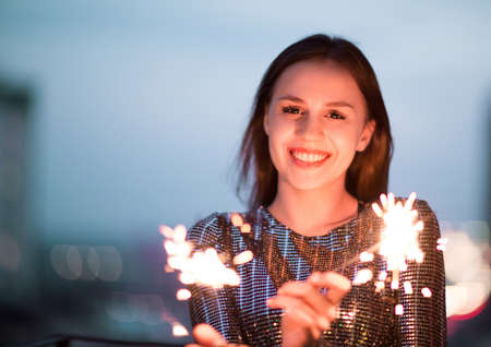 Delighted young lady in evening dress smiling and waving burning sparklers during holiday celebration at nightの写真素材
