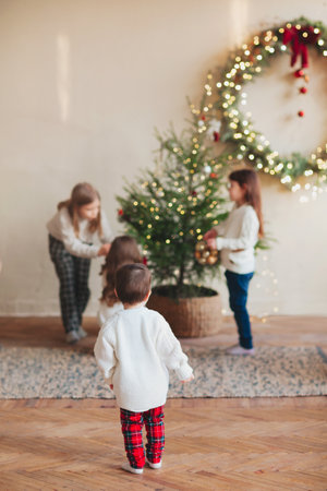 Happy four little siblings, three sisters and small brother in warm knitted sweaters gathered around fir-tree in living room and decorating it with toys and glossy balls. Family and christmas conceptの写真素材