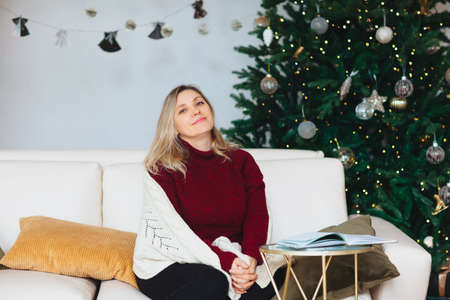 Happy caucasian joyful 50s woman sitting covered with warm knitted blanket on white cozy sofa on cushions with open book in her hands in modern living room decorated with sparking christmas fir-treeの写真素材