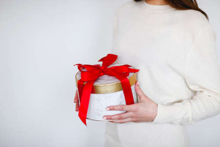 Unrecognizable girl in white sweater holding round present box tied with bright red ribbon during holiday celebrationの写真素材