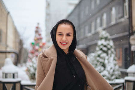 Young positive woman with funny smile on her face stands outside in beige coat and black scarf on head, shivering from winter cold weather against background of trees covered with thick layer of snowの写真素材