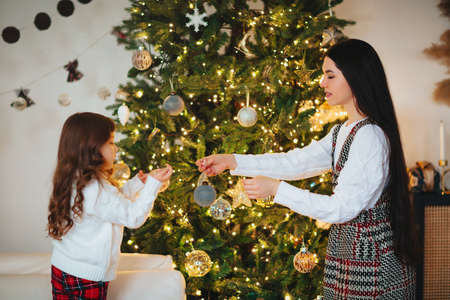 Joyful mother and lovely girl daughter in cozy knitted sweaters decorating Christmasmas fir tree with new year baubles and toys in living room, smile feeling atmosphere of winter holiday. Family holidaysの写真素材