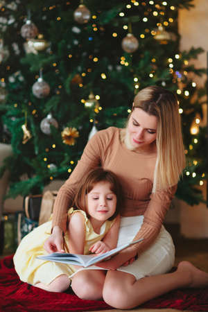 Happy family grandmother and little girl daughter sitting together on sofa and reading book while spending time together at home during Christmas holidays in winter, hugging and covering with wool plaidの写真素材