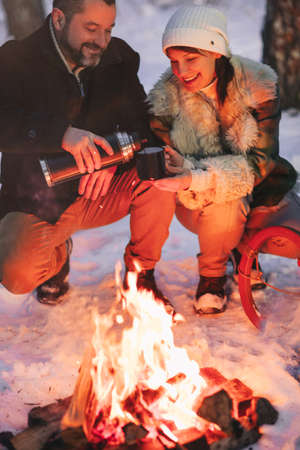 Joyful happy family couple in love basking by campfire outside in winter snowy forest drinking hot tea together, man and woman warm themselves hugging and loving look at each other. Trekking, adventure and seasonal vacationの写真素材