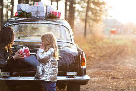 Cheerful young female with smiling daughter embracing against vintage black car with Christmas decoration and gift boxes with forest in backgroundの写真素材