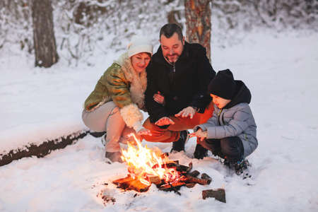 Happy family of three, mother, father and son, in warm clothes, sitting close to each other and trying to warm up by fire outside in cold snowy winter weather, against snowbound forest backgroundの写真素材