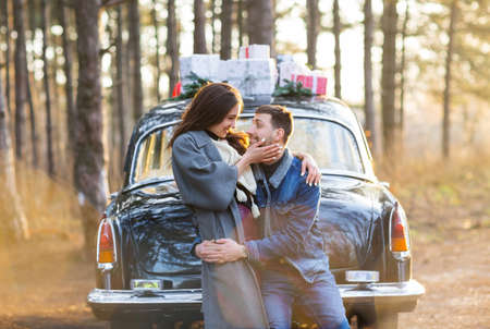 Side view of adult boyfriend and joyful woman in warm clothes embracing and looking at each other with love against black retro car in autumn forest at sunsetの写真素材