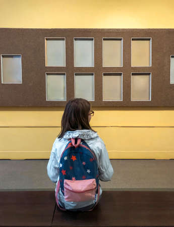Back view of teen girl with backpack in museum visiting exhibition, schoolgirl sitting on bench in art gallery and looking with interest at art objects or exhibits on wallの写真素材