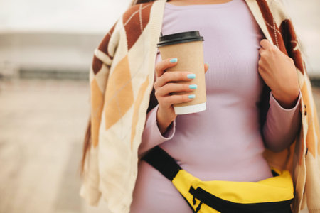 Cropped photo of pleased young woman with coffee to go cup standing outdoors, happy female wearing warm sweater holding hot drink and enjoying nice walk alone on autumn dayの写真素材