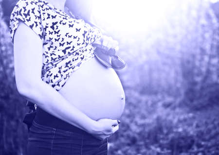Close up of unrecognizable pregnant woman with baby little shoes on her tummy at sunrise background. Color of 2022 year, very peri, tonedの写真素材