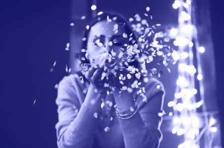 Happy beautiful brunette girl wearing sweater blowing on confetti isolated on vintage background with lights. Color of 2022 year, very peri, tonedの写真素材