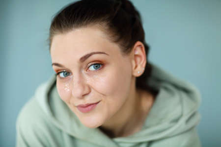 Beautiful portrait of young happy cheerful 30s woman in casual sweatshirt looking straight at camera smiling broadly and happy, showing her shiny glitter makeup, posing against gray wall backgroundの写真素材