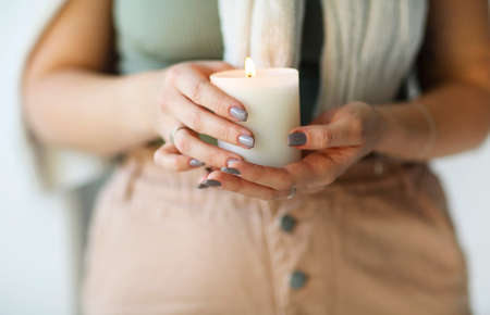 Side portrait of young calm woman holding lit candle in hands, closing eyes bringing close to her...