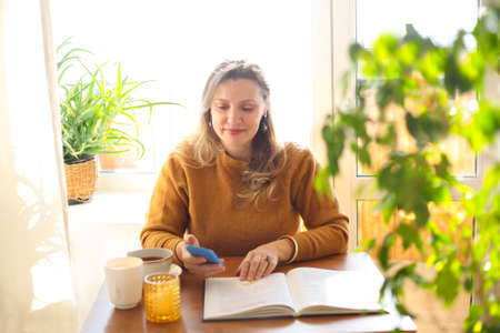 Smiling middle-aged woman looking at smartphone screen while sitting at table with open book and...