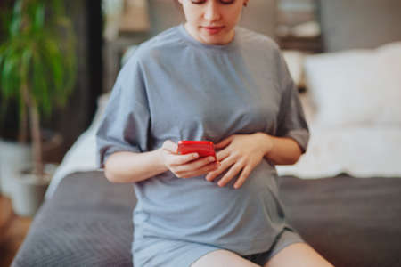 Young smiling pregnant woman touching belly talking on mobile phone with friend while spending time...
