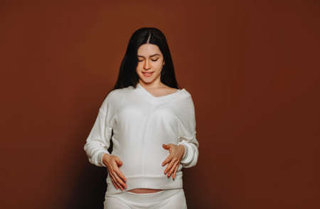 Young happy pregnant woman with long brown hair touching belly, talking with unborn baby while...