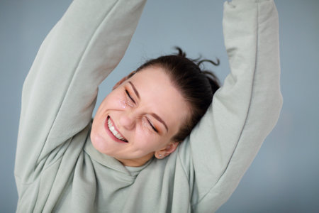 Beautiful portrait of young happy cheerful 30s woman in casual sweatshirt looking straight at camera smiling broadly and happy, showing her shiny glitter makeup, posing against gray wall backgroundの写真素材
