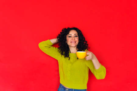 Young happy curly woman holding cup of tea, posing isolated on red wall. Smiling satisfied female enjoying hot drink, drinking coffee from mug while standing against bright studio backgroundの写真素材