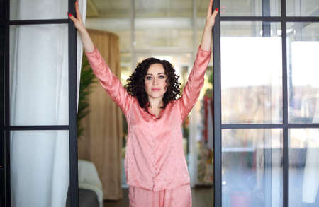 good morning. Positive indoor portrait of young attractive woman in silk pajama smiling at camera,...