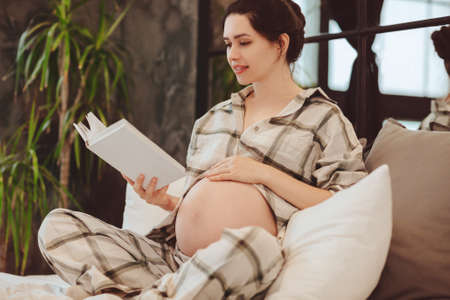 Young attractive happy pregnant woman in homewear reading book, touching belly while relaxing resting on couch at home, expectant mother wearing checkered pajama enjoying leisure time during pregnancyの写真素材